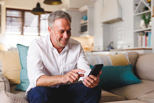 Happy caucasian man sitting on sofa in living room, using smartphone
