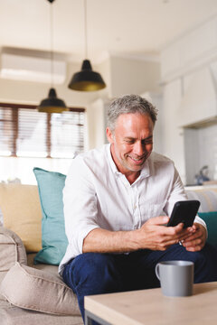 Happy caucasian man sitting on sofa in living room, using smartphone