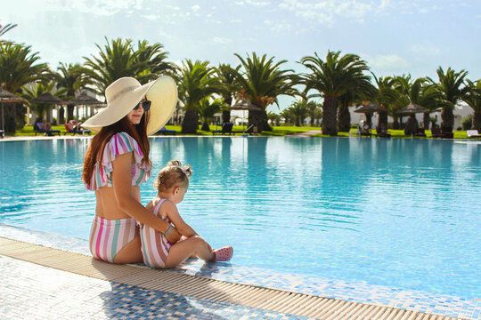 Young mom with little daughter sit near the basin with palm trees in identical striped pink swimsuits in the summer.