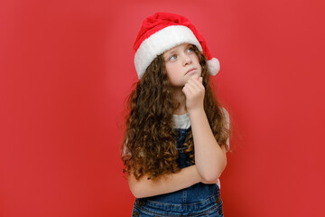 Portrait of pensive little girl child in Christmas hat holding hands on chin, posing isolated over pastel red color background wall in studio with copy space. Happy New Year 2023 holiday concept
