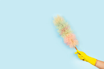 Mano con guante de látex amarillo sosteniendo un plumero de colores sobre un fondo celeste liso y aislado. Vista de frente y de cerca. Copy space - obrazy, fototapety, plakaty