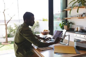 Happy african american man sitting at table in kitchen using laptop and tablet