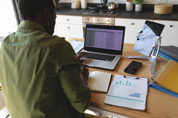 Happy african american man sitting at table in kitchen using laptop