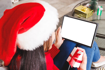 Biracial woman wearing santa hat, sitting on sofa in living room, using tablet with copyspace