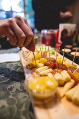 Hand taking a mini corn from a wooden board with salami, crackers, charcuterie, variety of cheeses, cherry tomatoes, nuts, etc