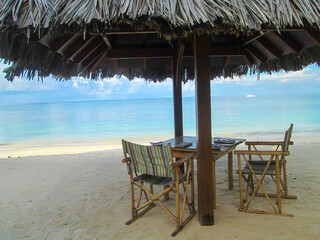 POV from a Tropical resort with no people on the beach and sun umbrella over a table with chairs,  horizon over water in a cloudy sky