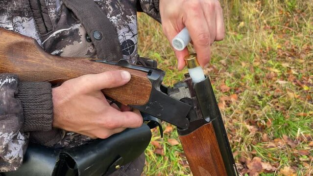 Close-up of a hunter in camouflage protective uniform, who is loading cartridges into a double-barreled shotgun.