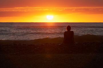A picturesque poppy sunset over the sea. A woman is sitting on the shore and admiring the setting sun. Selective focus.
