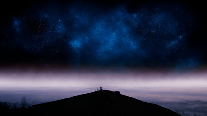 Fototapeta na wymiar A mystical concept of a man standing on a hill with dogs. Silhouetted against a night sky of a universe of stars. With a misty landscape below.