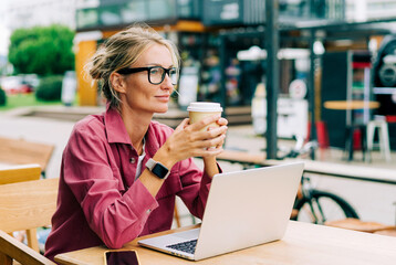 Smart modern woman sitting in a coffee shop drinking coffee and working on a laptop remotely.