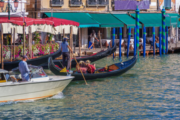A gondolier standing in a gondola steers a boat with an oar in his hands, an elderly couple of...