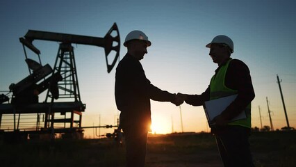 handshake oil contract. handshake a worker and businessman shaking hands against the backdrop of an oil pump. oil extraction business concept. silhouette sunlight handshake business contract - Powered by Adobe