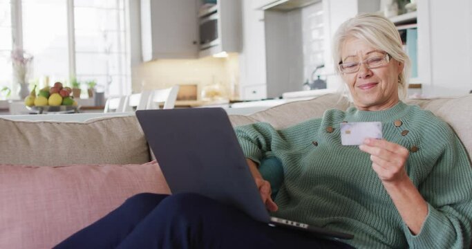 Happy senior caucasian woman sitting on sofa in living room, using laptop and holding credit card