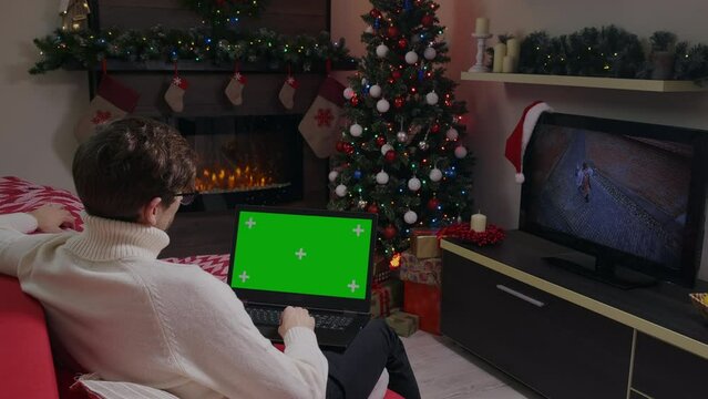 Man having video call on Green Screen Chroma key Mock up laptop. Girl video on background on TV. Winter season holiday edition.