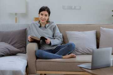 Happy Asian young woman wearing headphones with a book sitting relaxing on sofa at home