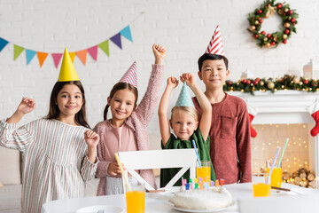 Excited multiethnic children in party caps looking at camera near birthday cake during party at home in winter