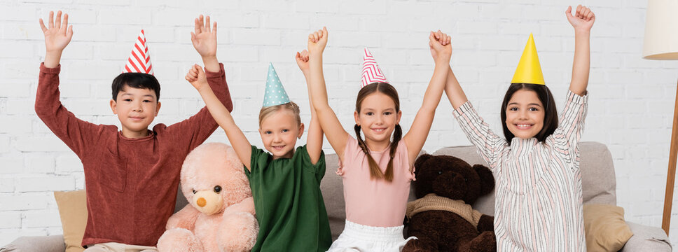 Cheerful interracial kids in party caps looking at camera during birthday party at home, banner
