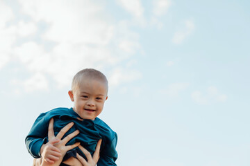 Cute blond boy with down syndrome on mom's arms, blue sky background