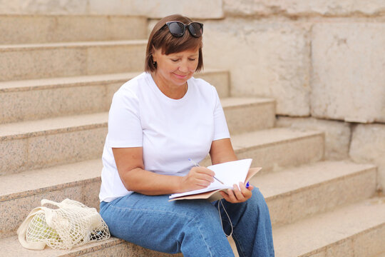 higher education for adults. mature woman in jeans and white t shirt writes and makes note in a notebook sitting on stairs outdoors, freelancer. College study program, academic educational course.