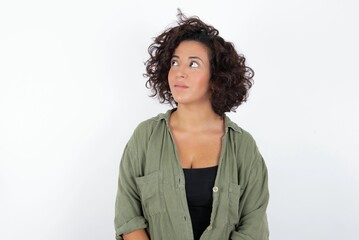 young beautiful woman with curly short hair wearing green overshirt over white wall looking aside into empty space thoughtful