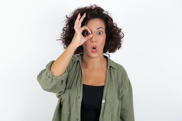 young beautiful woman with curly short hair wearing green overshirt over white wall doing ok gesture shocked with surprised face, eye looking through fingers. Unbelieving expression.