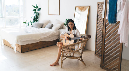 Brunette attractive female learning to play guitar while sitting in cozy home interior. Young concentrated woman enjoying her hobby with musical instrument performance relaxing on wicker chair