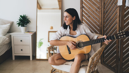 Brunette attractive female smiling and learning to play guitar while sitting in cozy home interior. Young happy woman enjoying her hobby with musical instrument performance relaxing on wicker chair