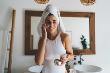 Young smiling female with towel on head puts moisturizing cream on face while standing at home bathroom. 30 years old happy woman doing daily morning rituals. Enjoying healthy skin with collagen care`