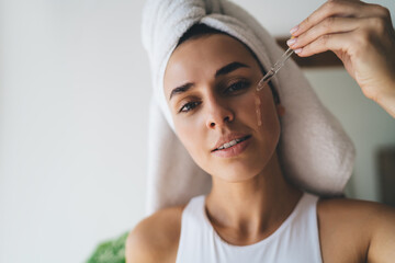 Young female with towel on head puts moisturizing serum on face while standing at bathroom. 30 years old woman doing daily morning rituals. Enjoying healthy skin with collagen care`, skincare product