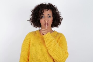 Surprised young beautiful woman with curly short hair wearing yellow sweater over white background makes silence gesture, keeps finger over lips and looks mysterious at camera