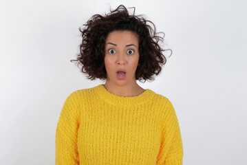 Shocked young beautiful woman with curly short hair wearing yellow sweater over white background stares bugged eyes keeps mouth opened has surprised expression. Omg concept