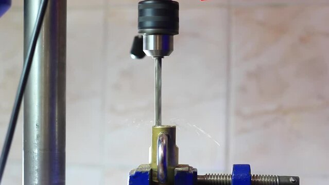 The drill drills a hole in a metal lock that is clamped with a vise