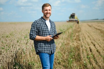 Handsome farmer with tablet standing in front of combine harvester during harvest in field.