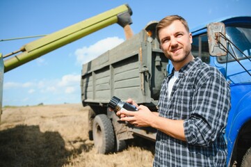 Happy farmer proudly standing in a field. Combine harvester driver going to crop rich wheat harvest. Agronomist wearing flannel shirt, looking at camera on a farmland