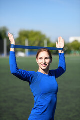 Women and sport. Smiling girl in sportswear - blue shirt and black leggings with a sports elastic band doing stretching on the grass at the stadium outdoor on a sunny day. Middle aged sportswoman