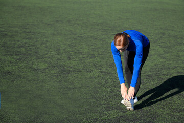 Women and sport. Girl in sportswear does exercises: bends and stretches on the grass at the stadium on a sunny day. Middle aged sportswoman dressed in sportsclothes exercising outdoors