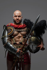 Portrait of isolated on grey background gladiator with muscular build holding helmet and spear.