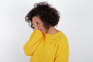 young beautiful woman with curly short hair wearing yellow sweater over white background with sad expression covering face with hands while crying. Depression concept.