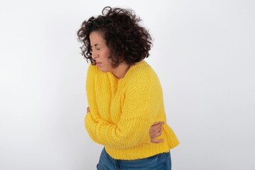 young beautiful woman with curly short hair wearing yellow sweater over white background got stomachache