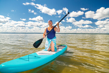 A woman in a pareo with a paddle on her knees on a SUP board in the lake against the background of white clouds on a clear blue sky.