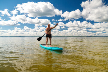 A man on a SUP board with a paddle in the lake against the background of white clouds on a clear blue sky.
