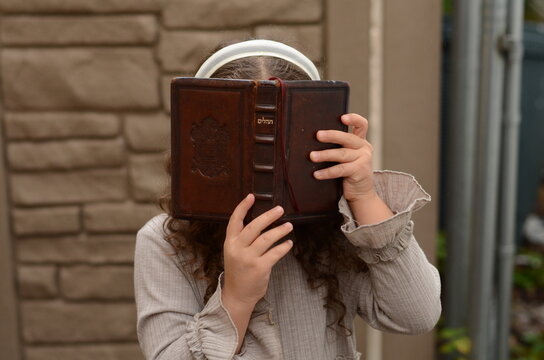 Little pretty Jewish girl with curly hair, wearing a dress and a white ribbon headband praying from a brown leather prayer book, tehillim, siddur outdoors