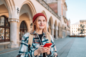 Happy blonde woman in hat using cellphone while walking at city street. High quality photo