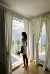 Bride in a dressing gown with a bouquet stands and looks out the window