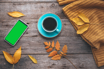 A cup of coffee and a mobile phone with a green screen on a wooden autumn background. Tea and autumn yellow leaves. Copy space.