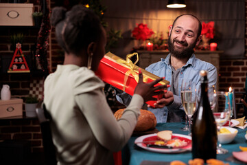 Obraz premium Couple celebrating christmas together, exchanging presents at festive table, eating traditional dishes at home feast. Man giving woman gift box, xmas celebration with friends
