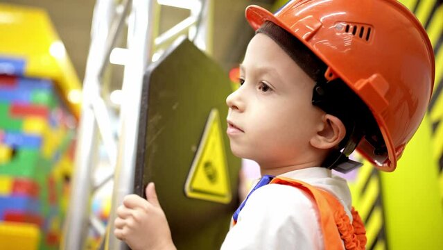 Baby boy stands in helmet and clothes of builder and makes house out of constructor bricks. Game in profession in children. Construction of toy building. Choosing future profession in childhood.