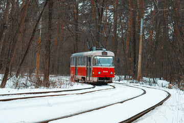 Old red tramway in the forest coming around the corner in winter with snow covered rails on foreground and naked trees on background - public transport and touristic attraction in Kyiv (Kiev), Ukraine