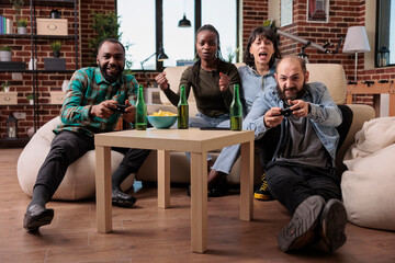 Happy friends laughing while they play strategy video games, using controller and tv console to have fun together. Young people enjoying leisure activity with beer bottles and gaming teamwork.
