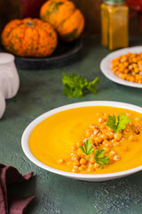 Creamy pumpkin soup with fried chickpeas garnish in a white ceramic plate on a green concrete background. Thanksgiving Day